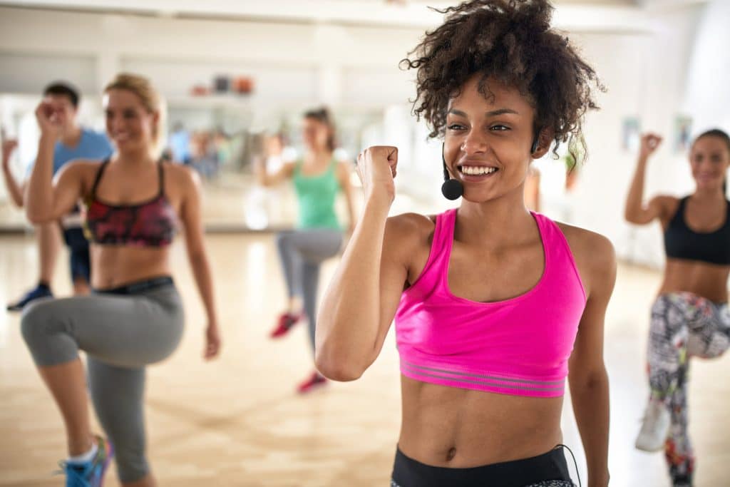 A lively group fitness class in progress, led by a woman in a pink sports top at the forefront, with participants in various athletic gear actively engaged in the background. The text 'Group Fitness Insurance' is prominently displayed, indicating the focus on insurance for group fitness activities.