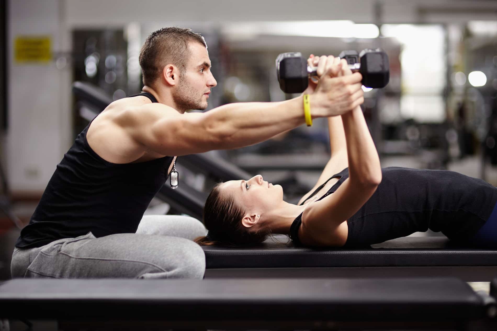 A personal trainer, a muscular man, is assisting a female client with bench presses using a dumbbell in a gym setting, exemplifying a scenario that would require personal trainer insurance for professional liability