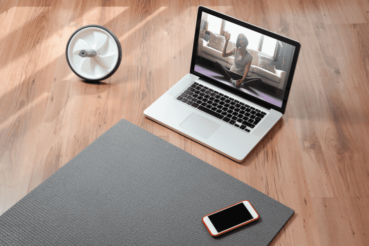 A laptop open on a wooden floor displays a fitness session, with a yoga mat, a smartwatch, and a speaker nearby, highlighting the context for Online Trainer Insurance.