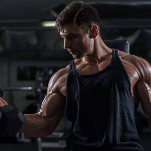 A muscular man lifting a dumbbell in a dimly lit gym, emphasizing the physical intensity of training and the potential risks that highlight the need for personal trainer insurance.