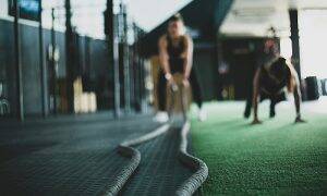 A person performing battle rope exercises in a gym with a trainer coaching in the background, symbolizing the steps to become a personal trainer in Pennsylvania.