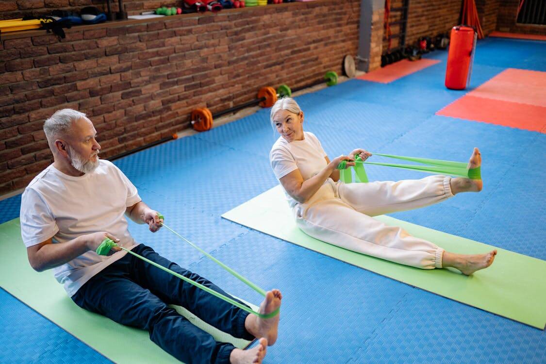 Senior man and woman using resistance bands for seated leg exercises during a fitness class tailored for older adults.