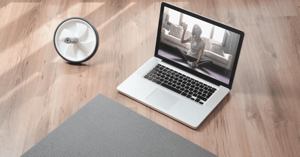 A laptop displaying a virtual workout session on a wooden floor, next to a yoga mat and an ab wheel, symbolizing the importance of liability waivers to protect personal training businesses.