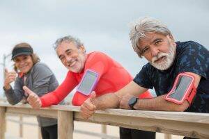 Three seniors smiling and giving thumbs up during an outdoor workout, wearing fitness gear and armbands for their phones.